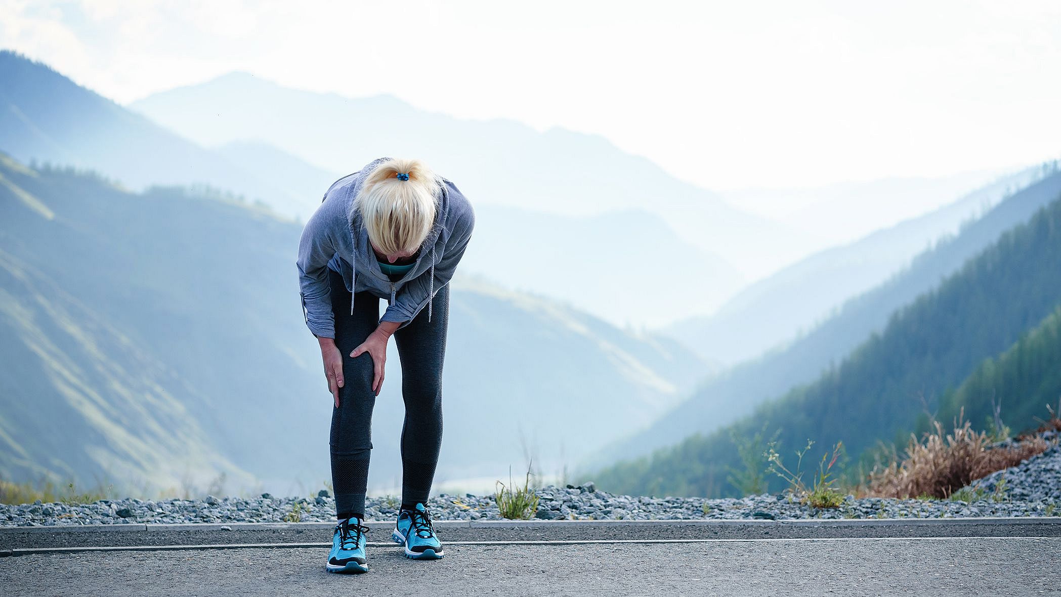 Die Aufnahme zeigt eine Joggerin. Sie steht auf einer Strasse, beugt sich nach vorne und hält ihre Hände auf das rechte Knie.